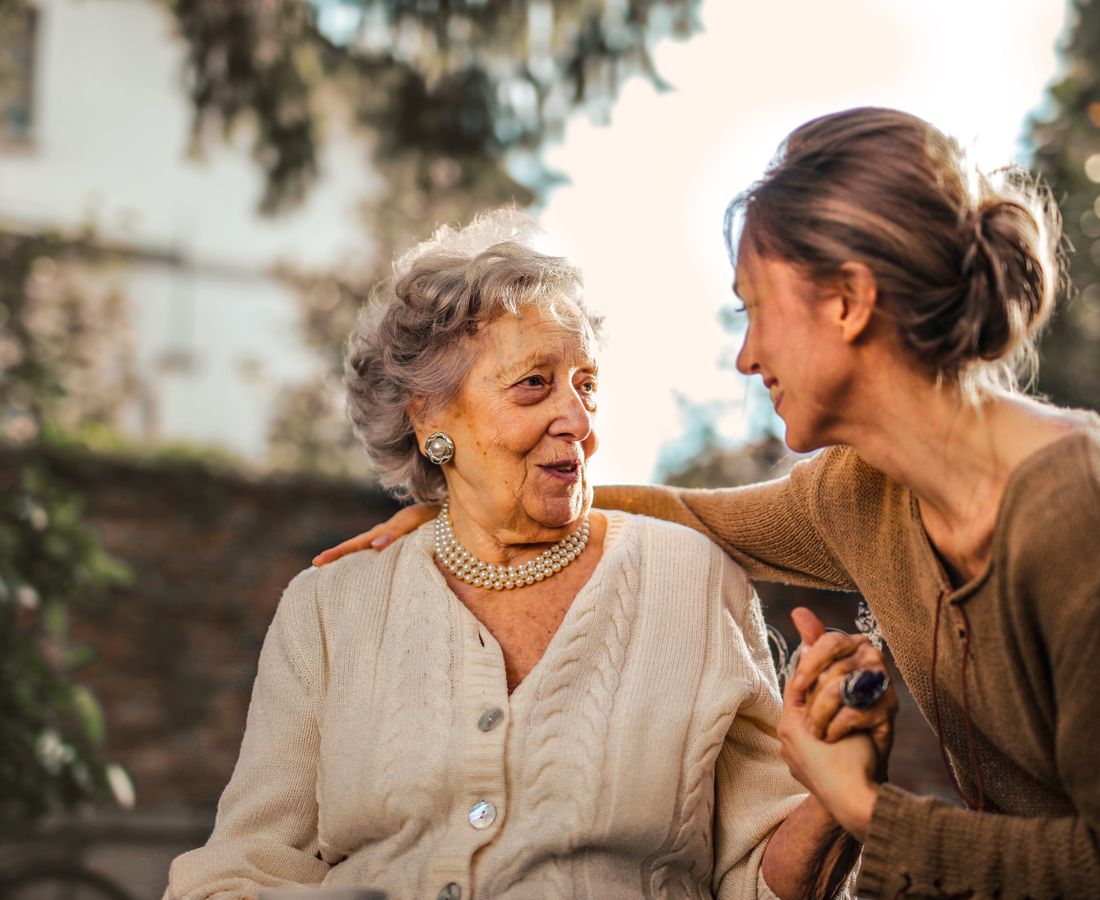 An adult daughter and her senior mother holding hands and smiling together in a sunny garden